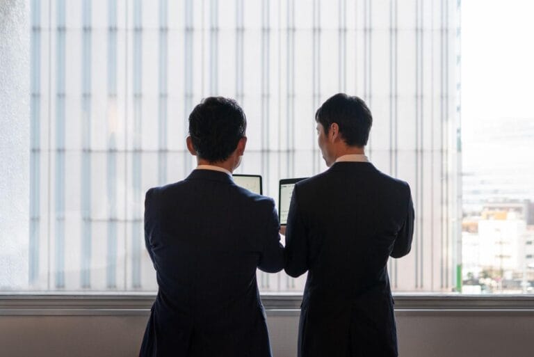 Cambodia Border Trade Reopening: Two business professionals in suits, standing by a window, examining information on laptops against a backdrop of a high-rise building.
